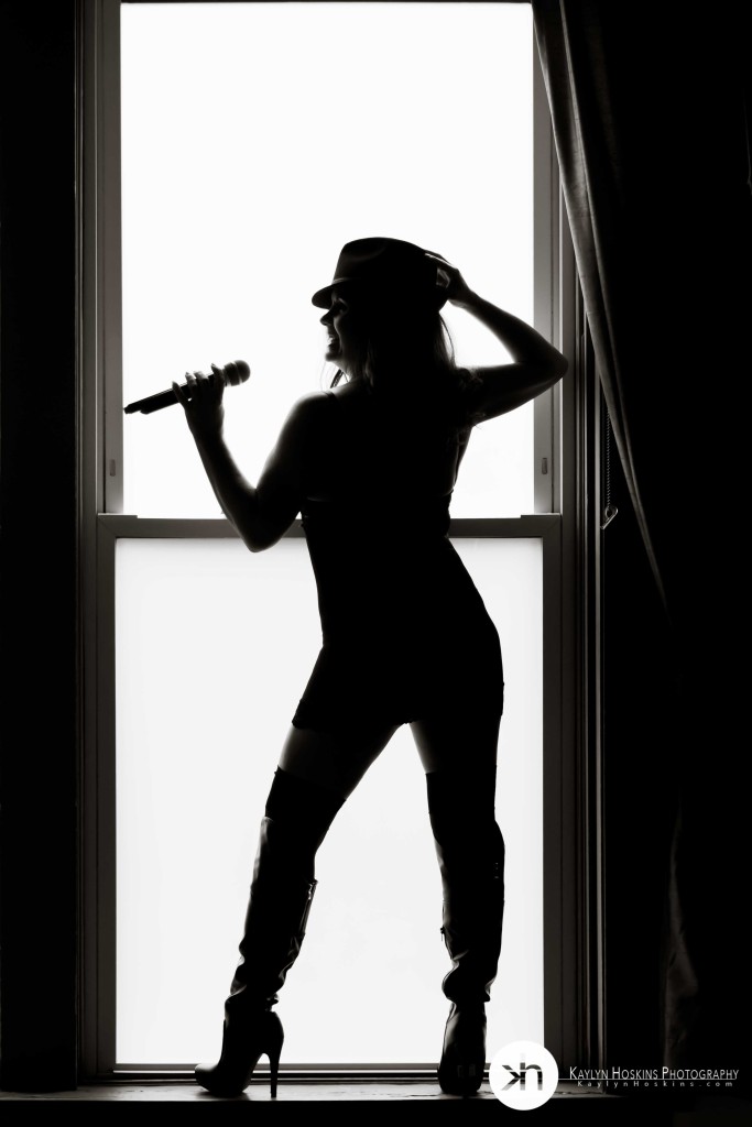 Boudoir client is a silhouette in large window holding microphone during luxury boudoir experience