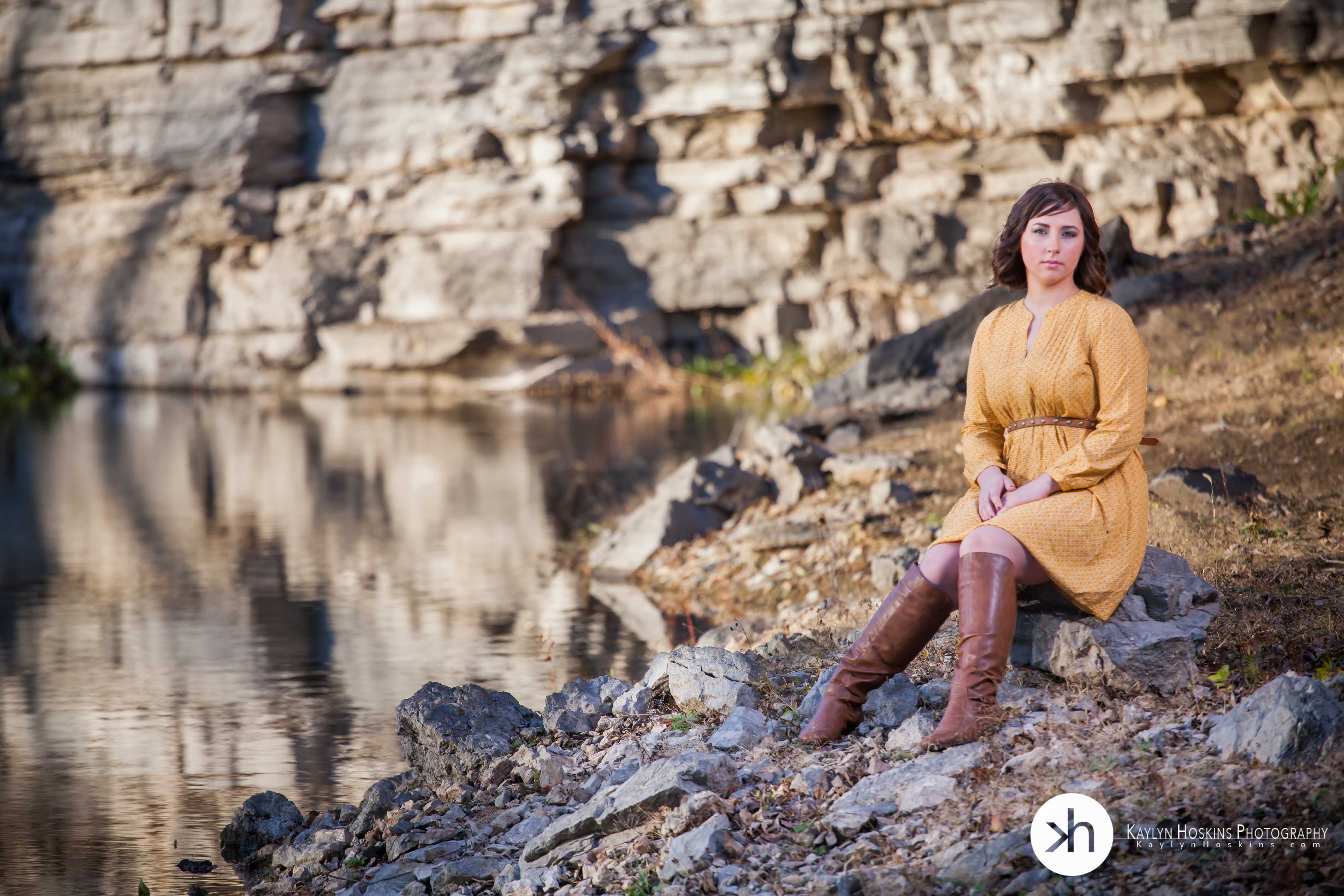 Madison Hamer sits on rock in mustard color dress by lake in north liberty, iowa