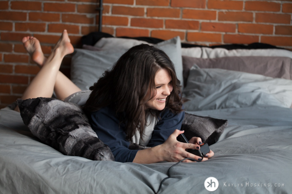 Boudoir Goddess laying on bed playing Atari during photo shoot at Kaylyn Hoskins Photography in Solon, IA  