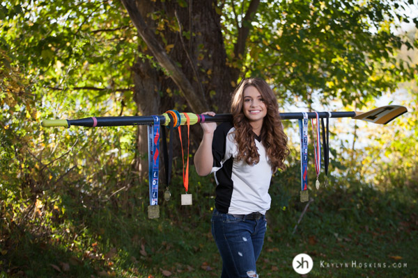 Solon Senior & Iowa Rower Aurora holds an oar on her shoulders with medals hanging on it during senior portrait session in iowa city