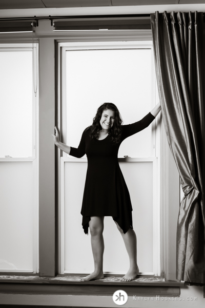 Prairie Senior wearing black dress in large window at Kaylyn Hoskins Photography studio in Solon