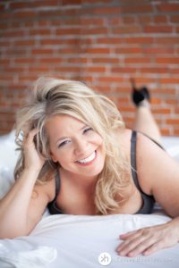 Gorgeous boudoir goddess smiles big at the camera while laying on bed in lingerie during boudoir photo shoot