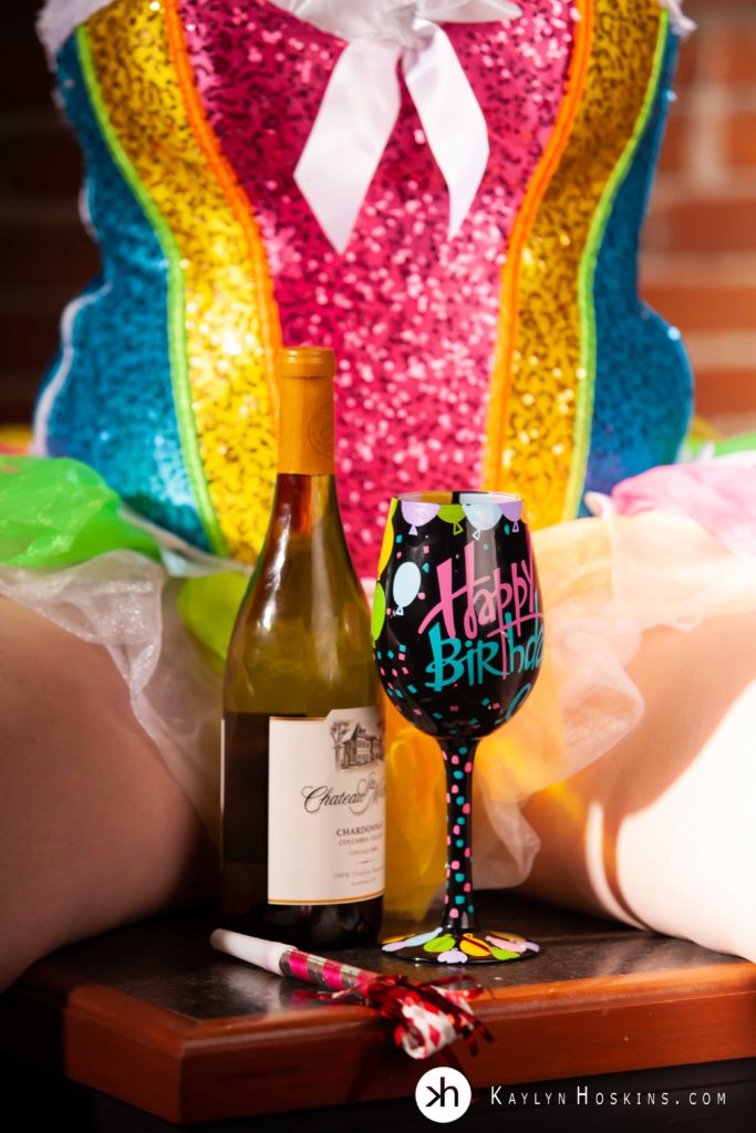 woman sitting on corner of bar in rainbow corset with tutu showing wine and happy birthday glass