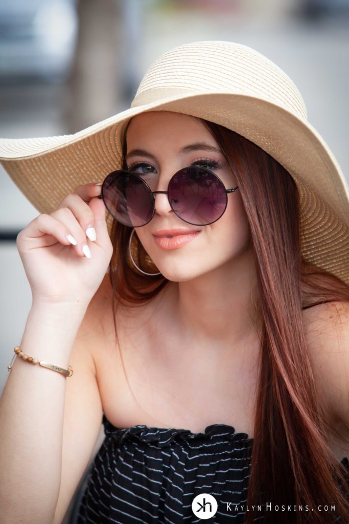 Jesup HS Senior peaks over large round hippie sunglasses wearing sun hat outside of Kaylyn Hoskins Photography downtown Solon, Iowa