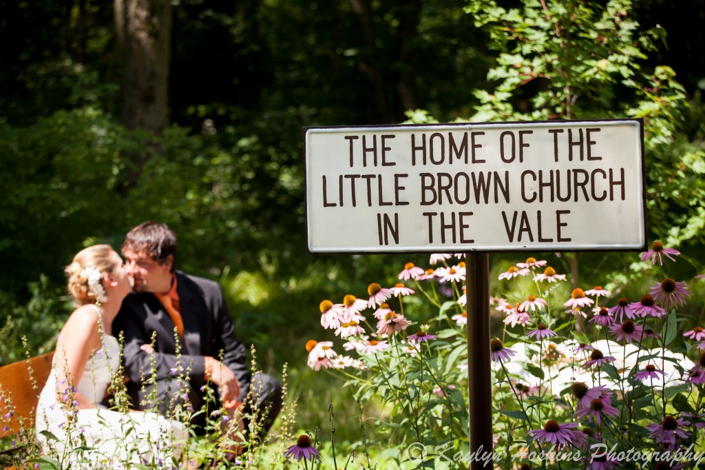 Bride and groom share a moment sitting on a bench around the beautiful flowers outside the Little Brown Church in Nashua, IA